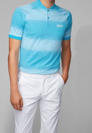 Homme portant un polo à manches courtes dégradé bleu avec le logo "BOSS" et un pantalon blanc, debout avec une main dans la poche.