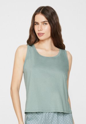 Femme aux longs cheveux bruns portant un débardeur vert clair sans manches et un pantalon vert clair à motifs sur un fond uni.
