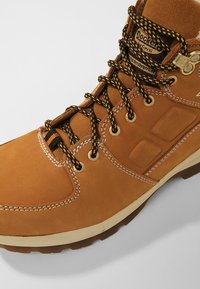 Bottine en suede marron avec des lacets noirs et jaunes, œillets dorés, col rembourré et surpiqûres blanches contrastantes. Semelle intermédiaire texturée.