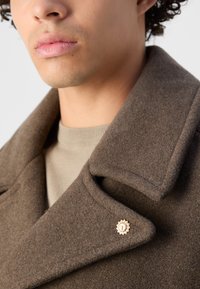 Brun ullblanding frakk med stor krage, med en dekorativ gullknapp. Beige strikkegenser under er synlig ved halslinjen.