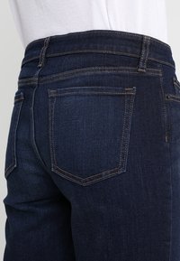 Tmavě modré džínové kalhoty s přiléhavým střihem, které mají standardní zadní kapsy a viditelné švy podél švů a pasu.