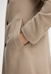 Beige wollen jas met een (notched) zak, vijf zwarte knopen en een zachte, textuurstof met een getailleerde pasvorm.