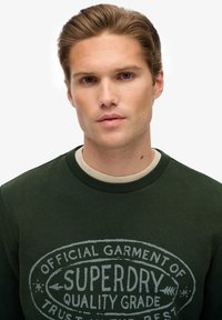 Jonge man met lichtbruin haar, die een donkergroene Superdry sweatshirt draagt, recht in de camera kijkt tegen een effen witte achtergrond.