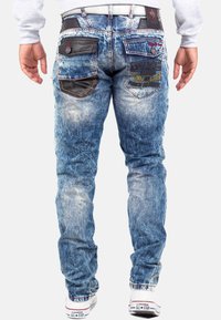 Jeans en denim bleu délavé avec une coupe ajustée, dotés de accents en cuir noir sur les poches et de coutures décoratives. Les poches arrière portent un détail textuel.