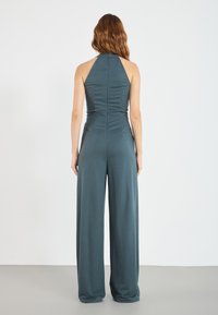Halter neck jumpsuit i dyp teal, laget av mykt stoff, med en tettsittende bodice og vide ben, med detaljer i baksemen.