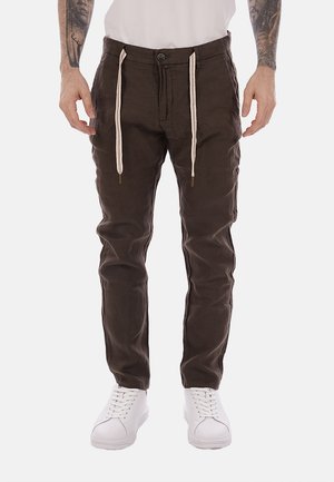 Hombre con pantalones ajustados de color marrón oscuro con cordones beige y zapatillas blancas, de pie con los brazos relajados a los lados.