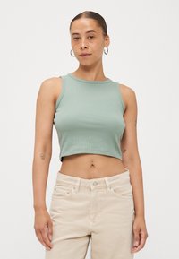 Zöld, bordázott crop tank top, kerek nyakkivágással. Bézs, magas derekú farmernadrággal párosítva, amely gombos és cipzáras záródással rendelkezik.