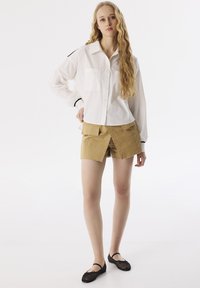 Camicia bianca con bottoni e maniche lunghe, con dettagli neri, abbinata a pantaloni corti color sabbia con tasche frontali e orlo grezzo. Scarpe nere basse.