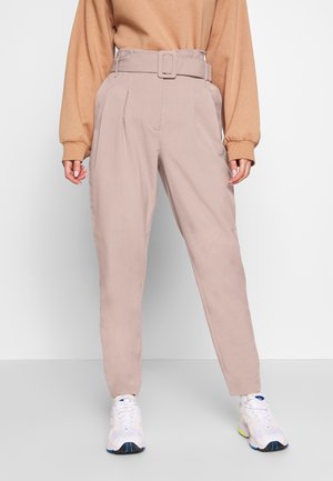 Pantalons taille haute rose clair avec jambes larges, détail de ceinture et plis avant, associés à des chaussures de sport blanches. Tissu texturé, design minimaliste.