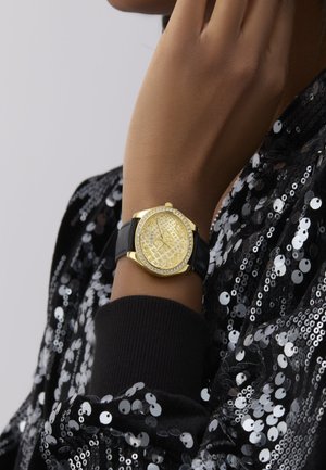 Žluté hodinky s texturovaným ciferníkem a akcenty z kamínků. Černý silikonový řemínek. Nosit se na ruce proti třpytivému černému oblečení posetému flitry.