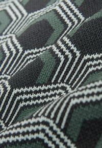 Gros plan d'un tissu tricoté avec un motif géométrique en noir, vert et blanc, comprenant des formes hexagonales et des lignes en zigzag.