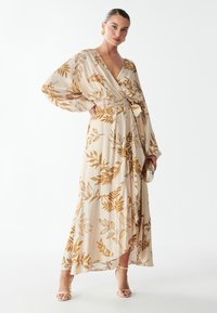Maxi-jurk in roomkleur met gouden loofdruk; heeft wijde mouwen, een wikkelontwerp en een zelfbindende ceintuur. Geaccessoireerd met een gouden clutch.