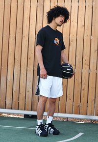 Camiseta negra de manga corta con diseño gráfico, pantalones cortos blancos y zapatillas negras. Sosteniendo un balón de baloncesto negro en una cancha al aire libre.