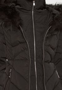 Sort polstret jakke med lynlås, der har en faux fur hætte og diagonalt quiltet mønster. Sølvhardware detaljer på lynlåsen.