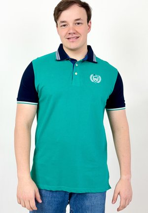 Jeune homme souriant, portant un polo à manches courtes bleu sarcelle et bleu marine avec un emblème, debout devant un fond blanc uni.