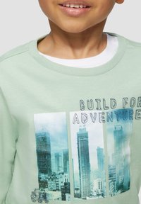 Hellgrüner Sweatshirt mit rundem Halsausschnitt, der eine fotografische Stadtszene-Grafik und den Text "BUILD FOR ADVENTURE" darüber zeigt.