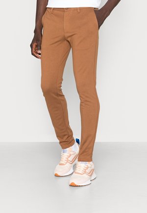 Man draagt slanke, geelbruine broek, witte sokken en beige sneakers met oranje details, staat met één hand in zak.