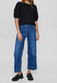 Blouse noire texturée à manches bouffantes, associée à un jean bleu à jambes larges et des chaussures noires épaisses, illustrant une tenue décontractée.