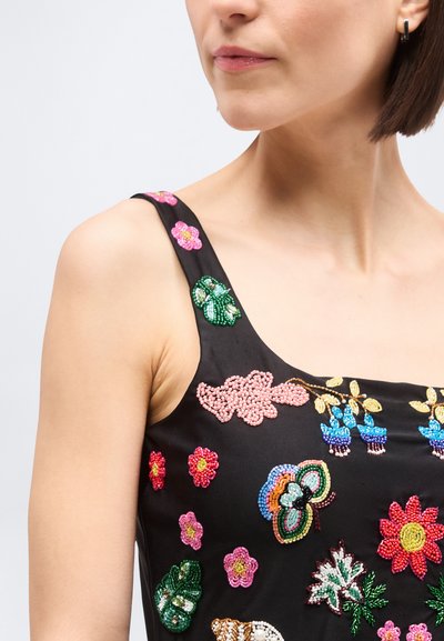 Kvinde iført sort ærmeløs top, pyntet med farverige perleblomster, en sommerfugl og grønne blade på brystet og skulderstroppen.