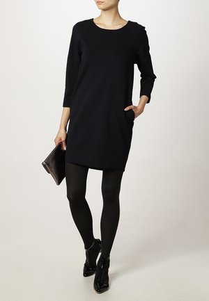 Robe noire mi-longue avec des manches trois-quarts, dotée d'un col rond et de poches latérales, portée avec des collants noirs et des chaussures à talons hauts élégantes.