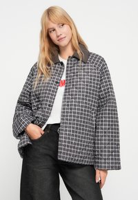 Πανωφόρι καπιτονέ σε γκρι και μαύρο καρώ σχέδιο με καφέ γιακά από κορντόροϊ. Διαθέτει χαλαρή εφαρμογή και κλεισίματα με κόπιτσες. Φοριέται με λευκό πουκάμισο.