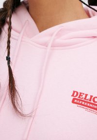 Sudadera de color rosa claro con cordones, que presenta un logo de texto en rojo que dice "DELICIOUS REFRESHING" en el pecho. Se puede apreciar la textura suave de la tela.