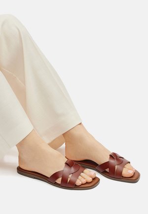 Braune Leder-Sandalen mit einem Kreuzriemen-Design, flacher Sohle und minimalistischem Stil, kombiniert mit lockeren cremefarbenen Hosen.