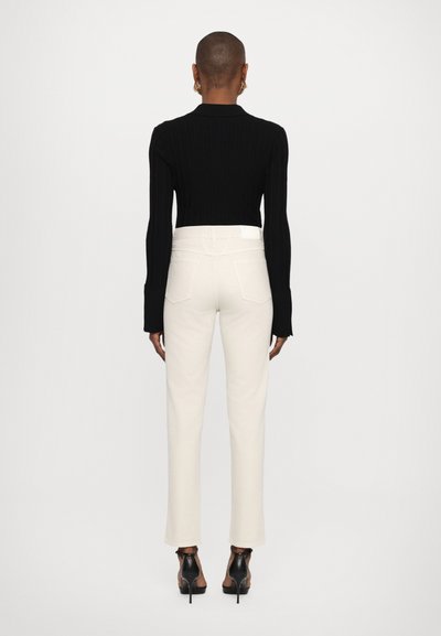 Black ribbed long-sleeve top paired with white straight-leg jeans. Model wearing black heeled sandals. Simple, modern design with minimal detailing.
