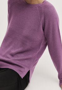 Lila gestrickter Pullover mit rundem Ausschnitt, gerippten Akzenten und seitlichen Schlitzern, kombiniert mit schwarzen Hosen. Weiche Textur und lockere Passform.