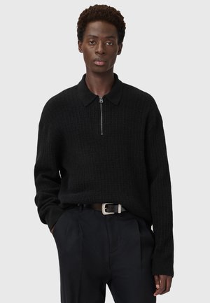 Pull noir côtelé avec une fermeture éclair, doté d'un col et de manches longues, associé à un pantalon sombre et une ceinture.