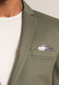 Blazer verde oliva con tessuto testurizzato, una tasca sul petto con un fazzoletto da taschino in grigio camouflage, indossato sopra una camicia bianca.