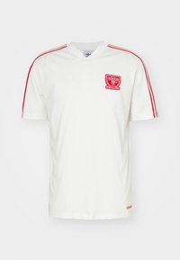 T-shirt branca de manga curta com três riscas vermelhas nos ombros e logótipo vermelho da Adidas bordado no peito. Design de gola em V, material em algodão.