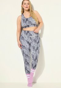 Mujer vestida con sujetador deportivo de tie-dye en tonos gris y negro, y leggings con calcetines a rayas rosas y blancos, de pie contra un fondo claro.