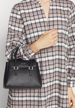 Person trägt ein beige-weiß kariertes Kleid und hält eine kleine schwarze Ledertasche mit zwei Griffen.
