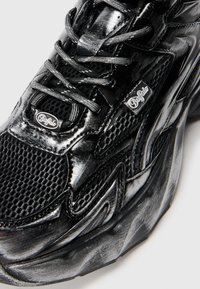 Sapatilha atlética preta com materiais sintéticos e malha brilhantes, parte superior texturizada, acentos de logótipos proeminentes e uma sola espessa e moldada.