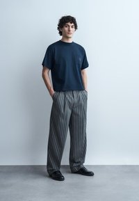 Młody mężczyzna stoi z rękami w kieszeniach, ubrany w granatowy T-shirt, luźne szare spodnie w prążki i czarne buty na jednolitym szarym tle.