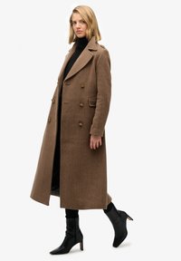 Brun uldfrakke med fiskegrøn mønster, store reverser, dobbeltradede knapper og sidelommer, kombineret med sorte ankelstøvler med høje hæle.