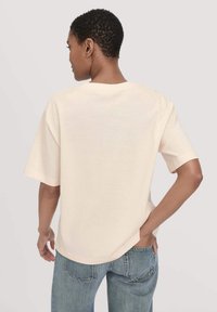 Person mit kurzen, lockigen Haaren, die ein lockeres, kurzärmliges beiges Shirt und blaue Jeans trägt, steht mit einer Hand in der Tasche, mit dem Rücken zur Betrachterin.