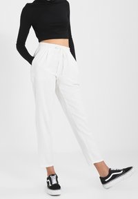 Pantalons blancs amples et fuselés avec une taille élastique. Portés avec un crop top noir à manches longues et des baskets noires.