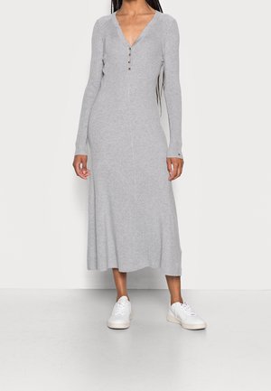 Kobieta w jasnoszarej, prążkowanej sukience midi z długim rękawem i guzikami na piersi, w połączeniu z białymi trampkami.