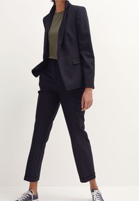 Granatowa marynarka o dopasowanym kroju z dwoma przednimi kieszeniami, zestawiona z dopasowaną oliwkową bluzką i ciemnymi spodniami, uzupełniona granatowymi sneakerami.