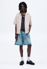 Kortærmet, beige tekstureret skjorte over en sort striktop, parret med lyseblå denim shorts. Tilbehør inkluderer en sølvhalskæde og sorte sko.