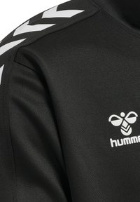 T-shirt de sport noir avec une texture lisse, présentant un logo blanc et un motif en chevrons sur les épaules. Conçu pour une utilisation athlétique.