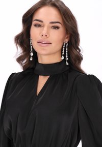 Abito in satin nero con colletto alto e scollatura a chiave, dotato di maniche lunghe con spalle arricciate. Accessoriato con orecchini a goccia in cristallo.