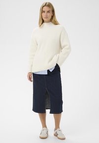 Femme portant un pull côtelé blanc sur une chemise rayée bleue, une jupe midi en denim foncé avec une fente à l'avant, et des baskets blanches avec des rayures noires.