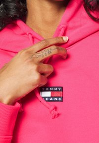 Un mano con un tatuaggio su un dito che tocca il petto di una felpa rosa brillante con un'etichetta del logo Tommy Jeans.