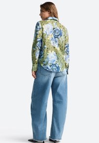 Floral long-sleeve shirt in green with blue and white flowers, featuring a collared neck. Paired with loose light blue denim jeans.