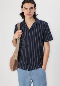 Chemise à manches courtes bleu marine avec des rayures verticales blanches, boutonnage à l'avant et texture douce, associée à un short en denim bleu et un sac beige.