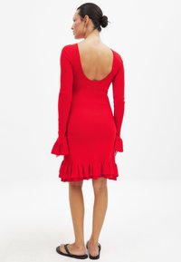 Rode geribbelde jurk met lange mouwen, open rug en uitlopende ruffle zoom. Soepele textuur en aansluitende pasvorm, gecombineerd met zwarte teenslippers.