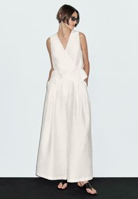 Witte linnen jumpsuit met een wikkelontwerp, brede benen en een ceintuur in de taille. Gestyled met zwarte zonnebrillen en sandalen. Soepele textuur.